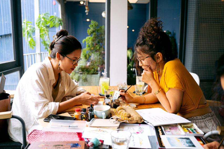 two women seated, leaning over table covered in art supplies