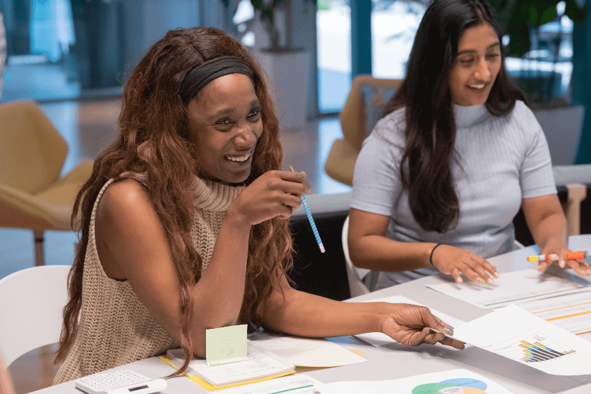 Two women sit at table covered with financial reports while smiling