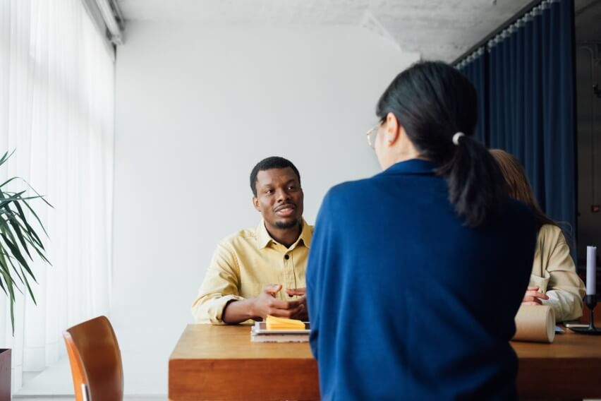 person with back to camera speaking to two other people across table in business setting